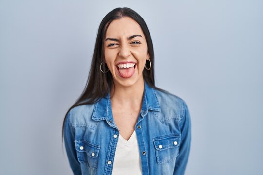 Hispanic woman standing over blue background sticking tongue out happy with funny expression. emotion concept.
