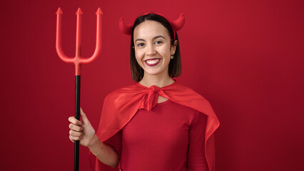 Young beautiful hispanic woman wearing devil costume holding trident over isolated red background
