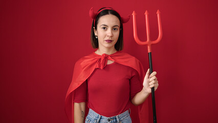 Young beautiful hispanic woman wearing devil costume holding trident over isolated red background