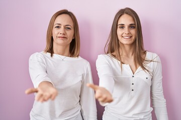 Middle age mother and young daughter standing over pink background smiling cheerful offering palm hand giving assistance and acceptance.