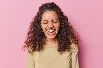 People and positive emotions concept. Happy woman with Afro hair bursts out from laughter keeps eyes closed giggles joyfully shows perfect white teeth dressed in casual brown t shirt pink backround