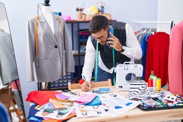 Young hispanic man tailor talking on smartphone writing on notebook at tailor shop