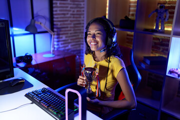 Fototapeta na wymiar Young african american woman streamer smiling confident holding trophy at gaming room