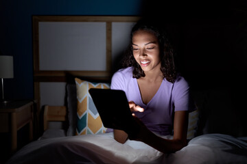 Young african american woman using touchpad sitting on bed at bedroom