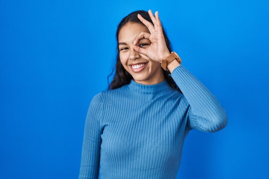 Young brazilian woman standing over blue isolated background doing ok gesture with hand smiling, eye looking through fingers with happy face.