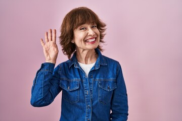 Middle age woman standing over pink background waiving saying hello happy and smiling, friendly welcome gesture