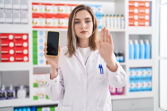 Blonde caucasian woman working at pharmacy drugstore showing smartphone screen with open hand doing stop sign with serious and confident expression, defense gesture
