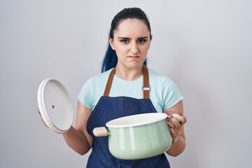 Young modern girl with blue hair wearing apron holding cooking pot clueless and confused...