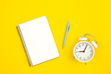 White alarm clock and blank notepad on yellow background, flat lay, copy space.
