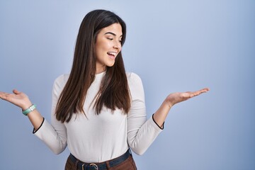 Young brunette woman standing over blue background smiling showing both hands open palms,...