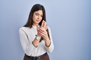 Young brunette woman standing over blue background suffering pain on hands and fingers, arthritis...