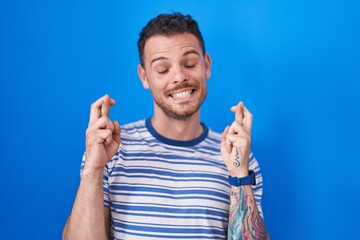 Fototapeta premium Young hispanic man standing over blue background gesturing finger crossed smiling with hope and eyes closed. luck and superstitious concept.