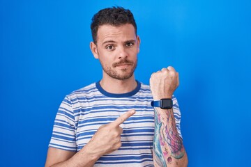 Young hispanic man standing over blue background in hurry pointing to watch time, impatience,...
