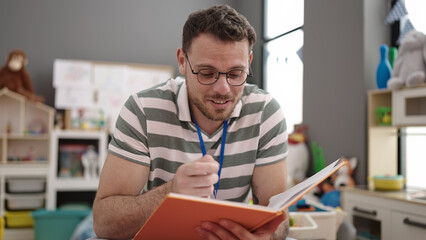 Young caucasian man working as teacher looking at notebook explaining at kindergarten