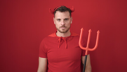 Young caucasian man standing with serious expression wearing devil costume over isolated red...