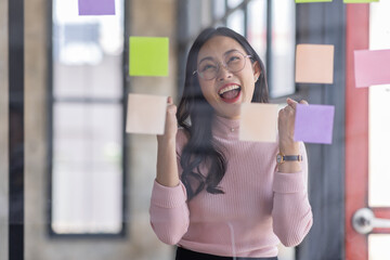 Obraz premium Young Serious asian Creative team use post it notes to share idea sticky note on glass wall. Asian business people design planning and Brainstorming thinking sticky History notes concept.