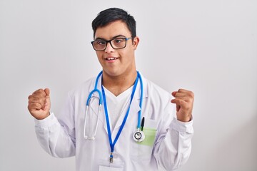 Young hispanic man with down syndrome wearing doctor uniform and stethoscope screaming proud, celebrating victory and success very excited with raised arms