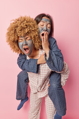 Sleepy two young women awake early in morning undergo beauty procedures apply facial mud mask dressed in comfortable nightwear give piggyback ride yawn and cover mouth isolated over pink background.