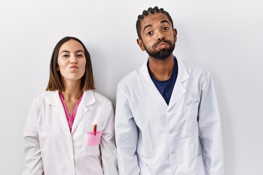 Young hispanic doctors standing over white background puffing cheeks with funny face. mouth inflated with air, crazy expression.