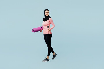 Full body young asian muslim fitness trainer sporty woman wear pink abaya hijab spend time in home gym walk hold yoga mat bottle water isolated on plain blue background studio Sport fit abs concept