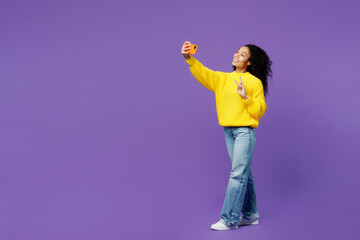 Full body young woman of African American ethnicity wear casual yellow sweater doing selfie shot on mobile cell phone post photo on social network isolated on plain purple background studio portrait.