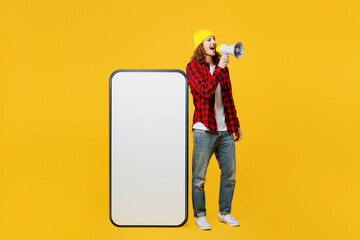 Full body young man wearing red checkered shirt white t-shirt hat big huge blank screen mobile cell phone smartphone with area scream in megaphone isolated on plain yellow background studio portrait.