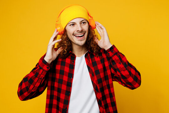Young smiling cheerful happy caucasian man wearing red checkered shirt white t-shirt hat headphones listen to music look aside isolated on plain yellow background studio portrait. Lifestyle concept.
