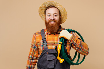 Young bearded man wear straw hat overalls work in garden hold hose with sprinkler on shoulder...