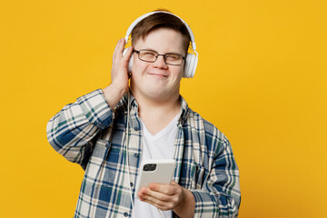 Young satisfied man with down syndrome wear glasses casual clothes headphones listen to music use mobile cell phone isolated on pastel plain yellow color background. Genetic disease world day concept.