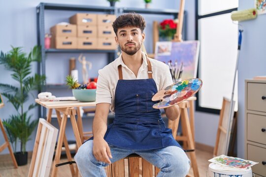 Arab man with beard painter sitting at art studio holding palette thinking attitude and sober expression looking self confident