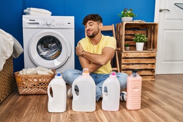 Arab man with beard doing laundry sitting on the floor with detergent bottle hugging oneself happy and positive, smiling confident. self love and self care