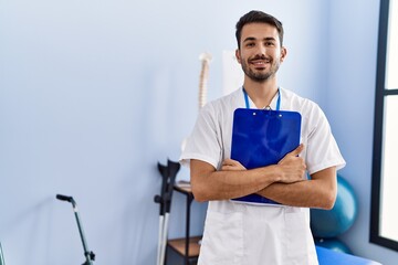 Young hispanic man wearing physiotherapist uniform holding clipboard at rehab clinic