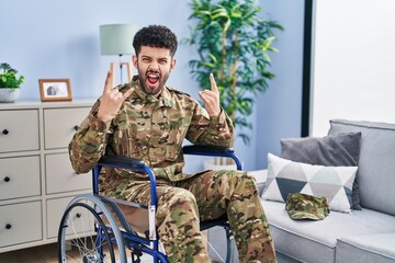 Arab man wearing camouflage army uniform sitting on wheelchair shouting with crazy expression doing rock symbol with hands up. music star. heavy concept.