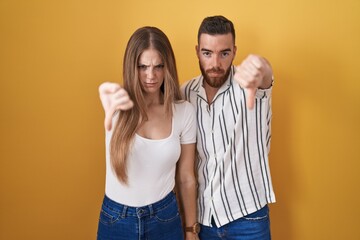 Young couple standing over yellow background looking unhappy and angry showing rejection and negative with thumbs down gesture. bad expression.