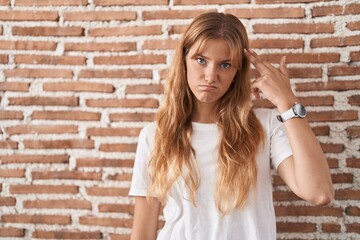 Young caucasian woman standing over bricks wall shooting and killing oneself pointing hand and fingers to head like gun, suicide gesture.
