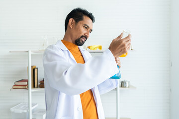 A male teacher in white lab coat with laboratory tools background in university science classroom checking at two blue and yellow chemical bottles for teaching class in both bare hands with carefully.