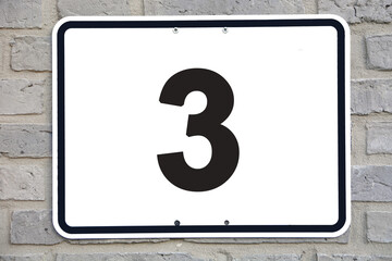 A white number plaque, on a grey brick wall, showing the number 3 (number three)