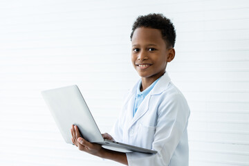 A little short hair African boy in white lab coat holding computer laptop in both hands. He smiles happily while preparing and checking data experiment for presentation teacher in chemistry classroom.