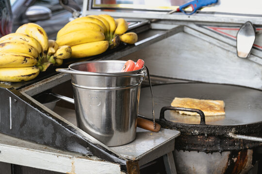Workplace for cooking strid food. A bucket with kitchen utensils, bananas and a large pan for making fried pancakes with bananas.
