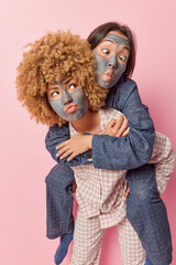 Two young women apply facial clay mask to reduce pores and fine lines dressed in pajama give piggyback ride foolish around after awakening isolated over pink background. Beauty procedures concept
