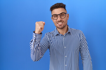 Hispanic man with beard wearing glasses angry and mad raising fist frustrated and furious while...