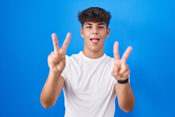 Hispanic teenager standing over blue background smiling with tongue out showing fingers of both hands doing victory sign. number two.