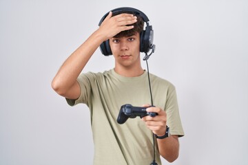 Hispanic teenager playing video game holding controller surprised with hand on head for mistake, remember error. forgot, bad memory concept.
