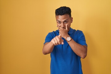 Young hispanic man standing over yellow background laughing at you, pointing finger to the camera with hand over mouth, shame expression