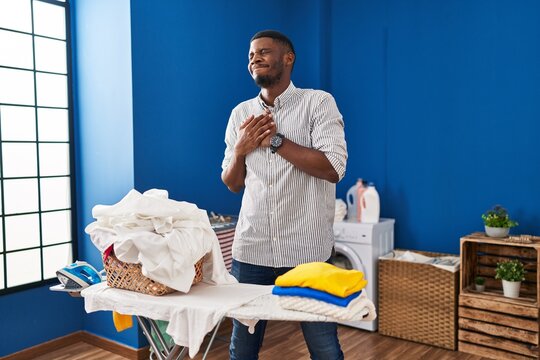 African american man ironing clothes at home smiling with hands on chest, eyes closed with grateful gesture on face. health concept.