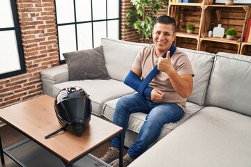 Obraz premium Hispanic young man wearing arm on sling for motorbike accident smiling happy and positive, thumb up doing excellent and approval sign