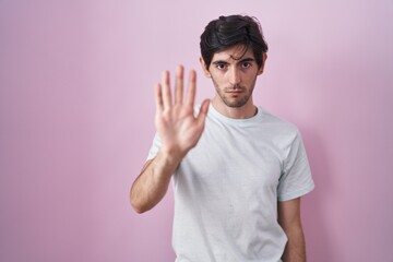 Young hispanic man standing over pink background doing stop sing with palm of the hand. warning expression with negative and serious gesture on the face.