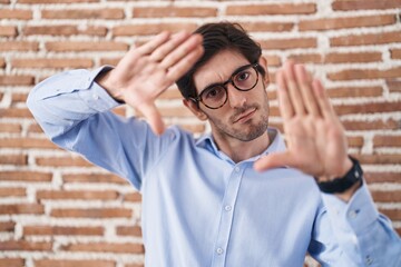 Young hispanic man standing over brick wall background doing frame using hands palms and fingers, camera perspective