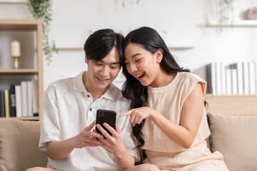 Asian young couple sit on couch or sofa looking at mobile phone smile and laughing together....