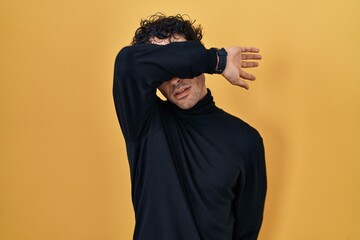 Hispanic man standing over yellow background covering eyes with arm, looking serious and sad....